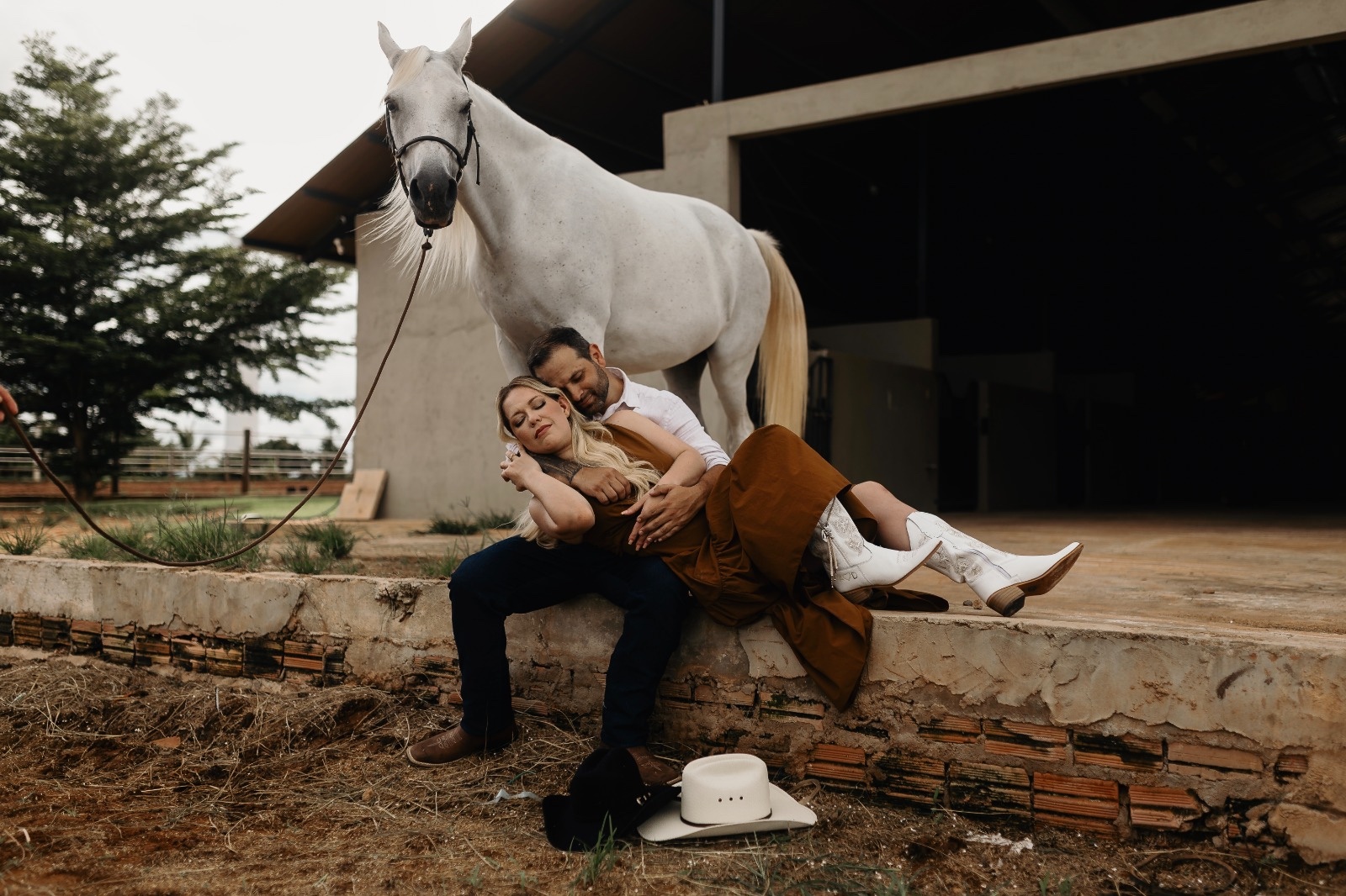Casal abraçado junto a um cavalo branco em frente ao haras